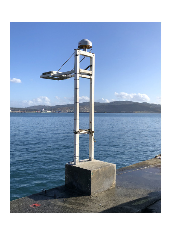 A GNSS antenna mounted on a frame above a concrete base at the edge of a quay, with the sea and hills behind, Langosteira, Spain.