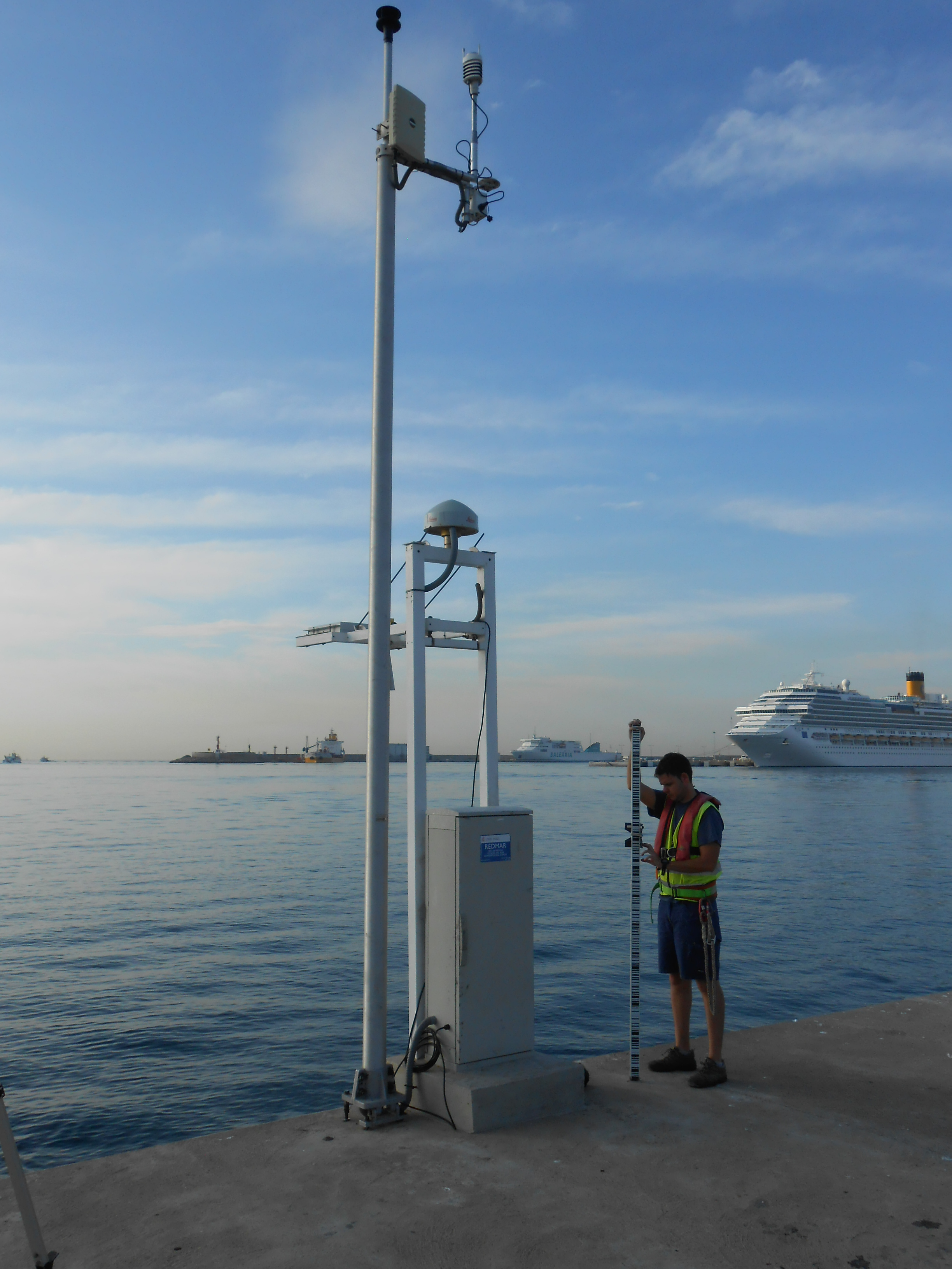 A REDMAR tide gauge station on a quay in the port of Palma, Mallorca, with a GNSS antenna and radar sensor, while a technician holds a levelling staff beside it.